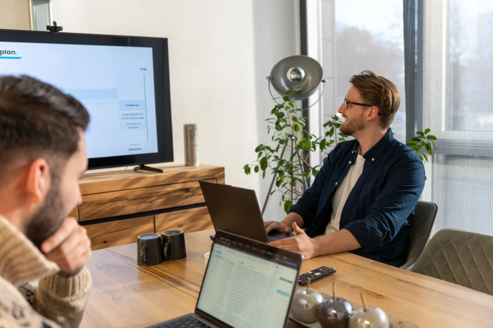 Zwei Männer sitzen an einem Holztisch mit Laptops, einer blickt auf einen großen Bildschirm mit Präsentation, im Hintergrund stehen eine Stehlampe und eine Pflanze.