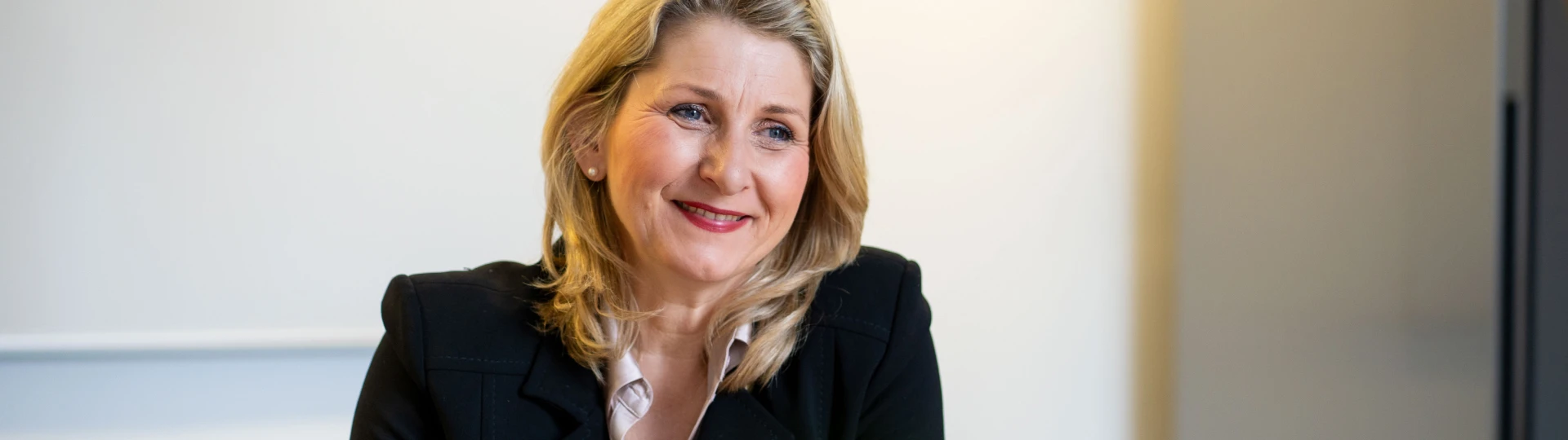 Frau mit blonden Haaren in schwarzem Blazer lächelnd vor hellem Hintergrund sitzend.