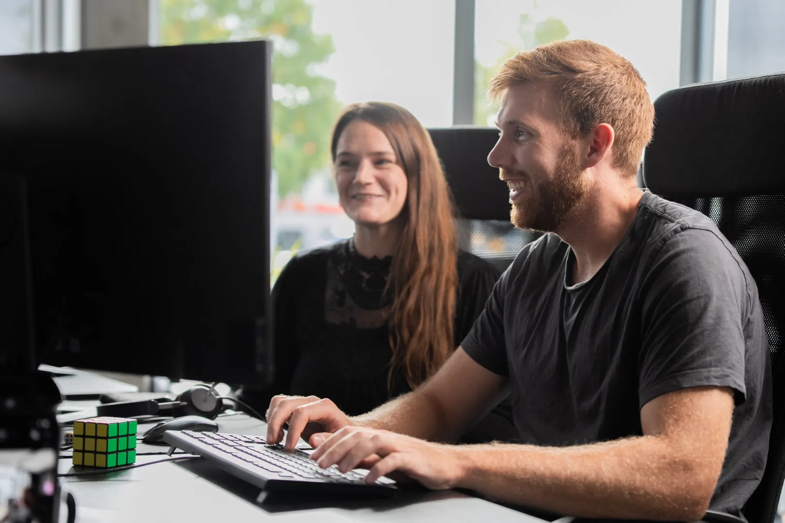 Ein Mann mit Bart sitzt lächelnd vor einem Computerbildschirm, daneben eine Frau mit langen Haaren, auf dem Tisch liegt ein Zauberwürfel.