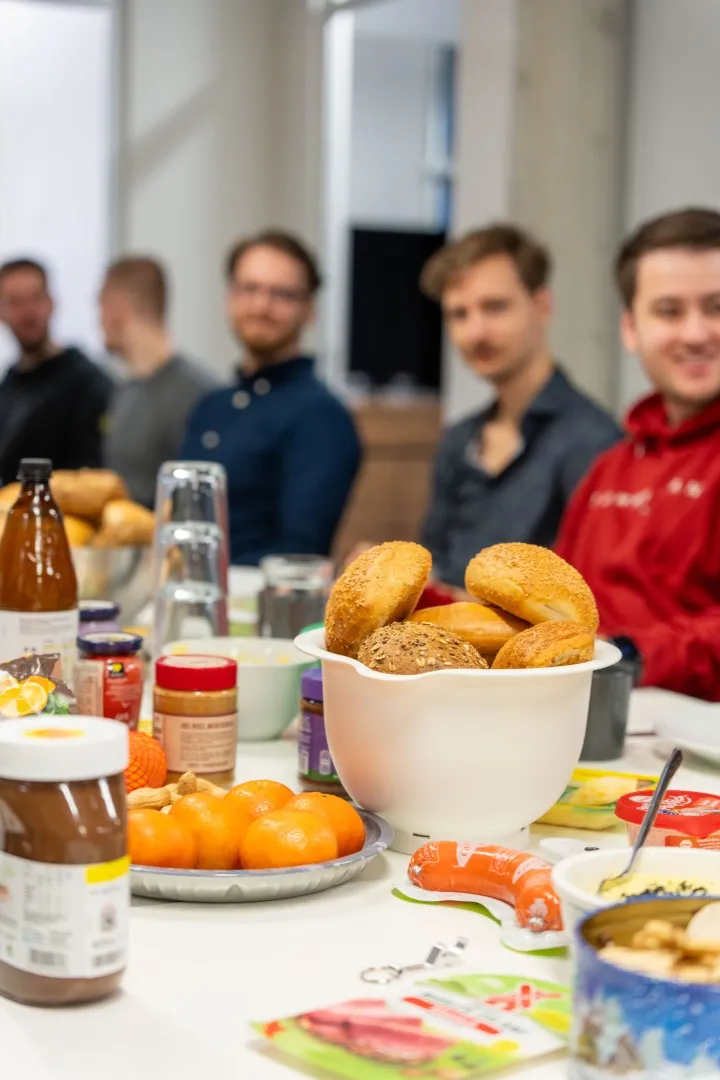 Reich gedeckter Frühstückstisch mit Brötchen und Aufstrichen, im Hintergrund sitzen mehreren Personen.
