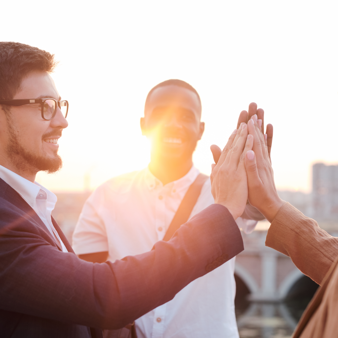 Drei Personen in Hemden und Anzügen stehen vor einem Sonnenuntergang und geben sich ein High Five. 