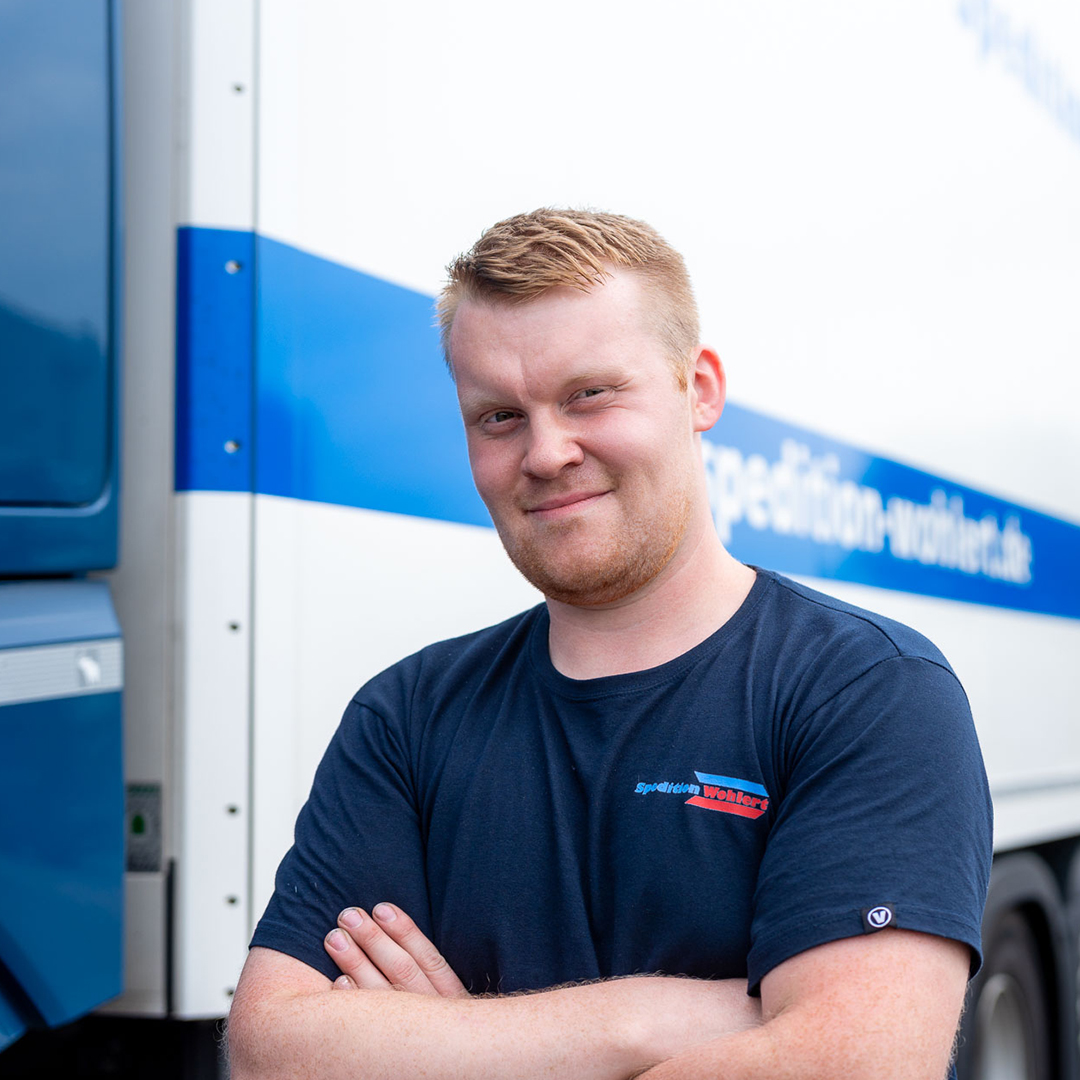 Ein LKW-Fahrer von Spedition Wohlert steht lächelnd mit verschränkten Armen vor einem LKW. 